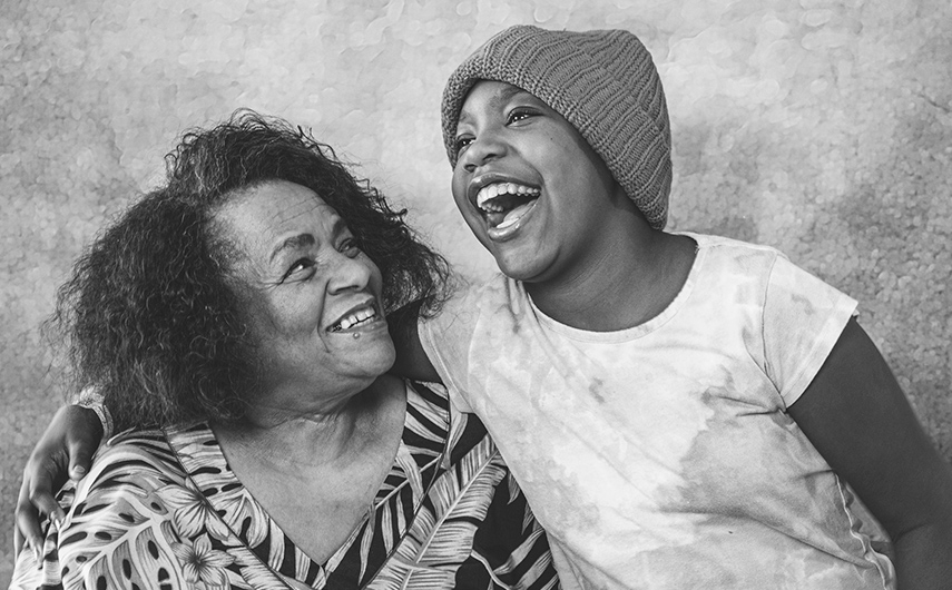 An older woman and a young girl wearing a knit cap smile and laugh together, with the girl’s arm around the woman’s shoulder.