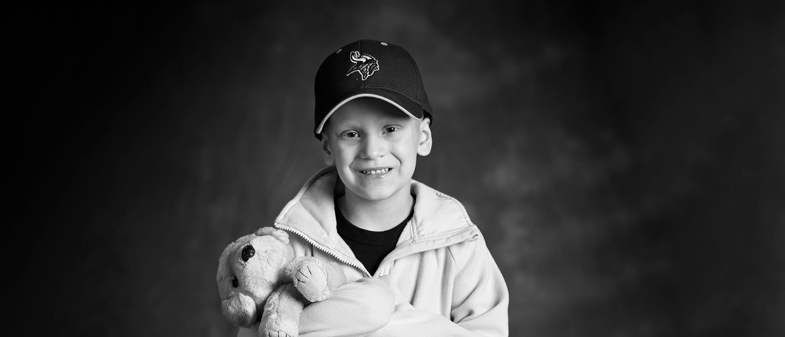 Black and white photo of a child with caner wearing a baseball cap and holding a stuffed animal